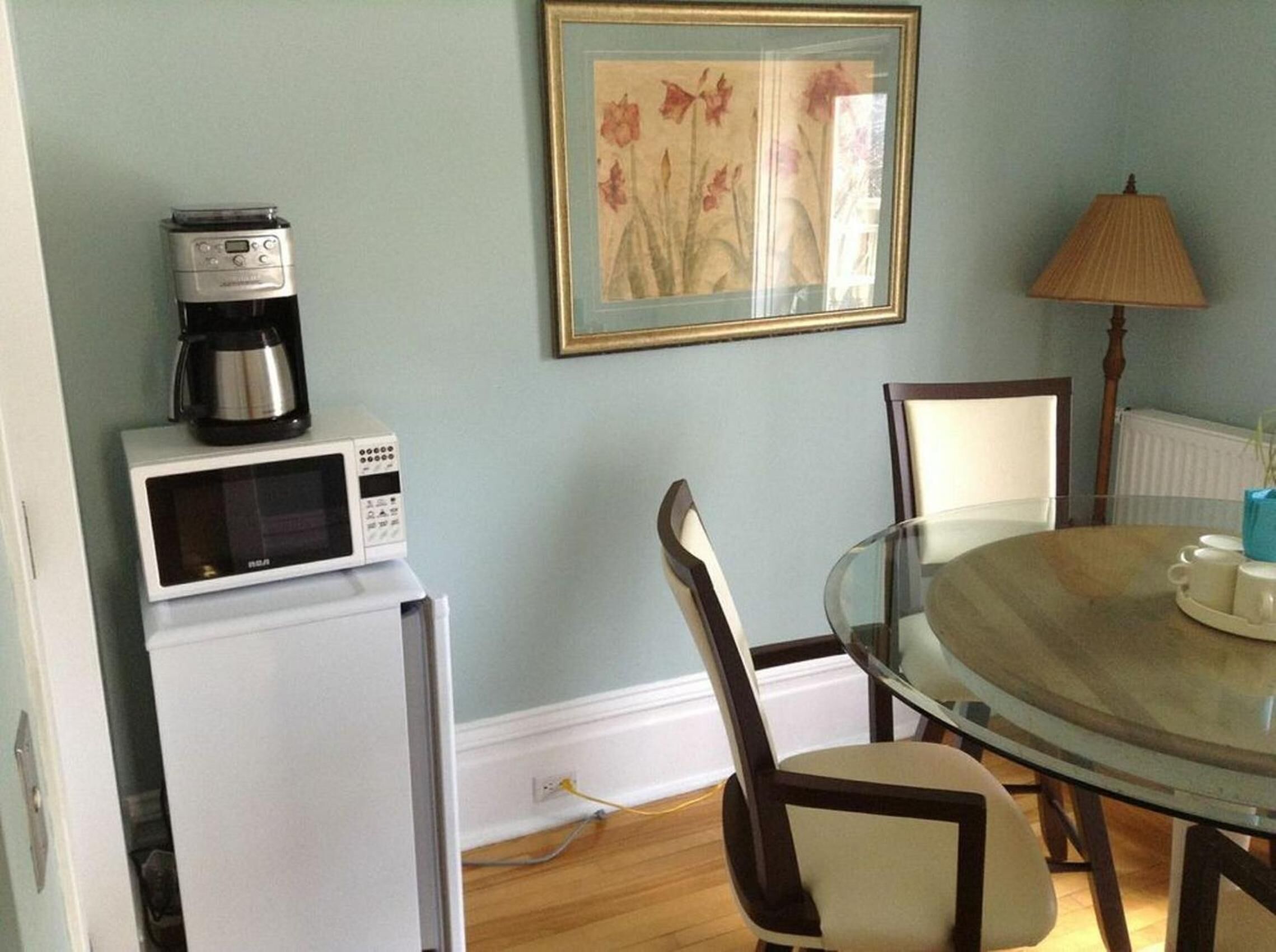 A dining area with a round glass table, white chairs, a coffee maker, a microwave, and a framed floral painting on the wall.