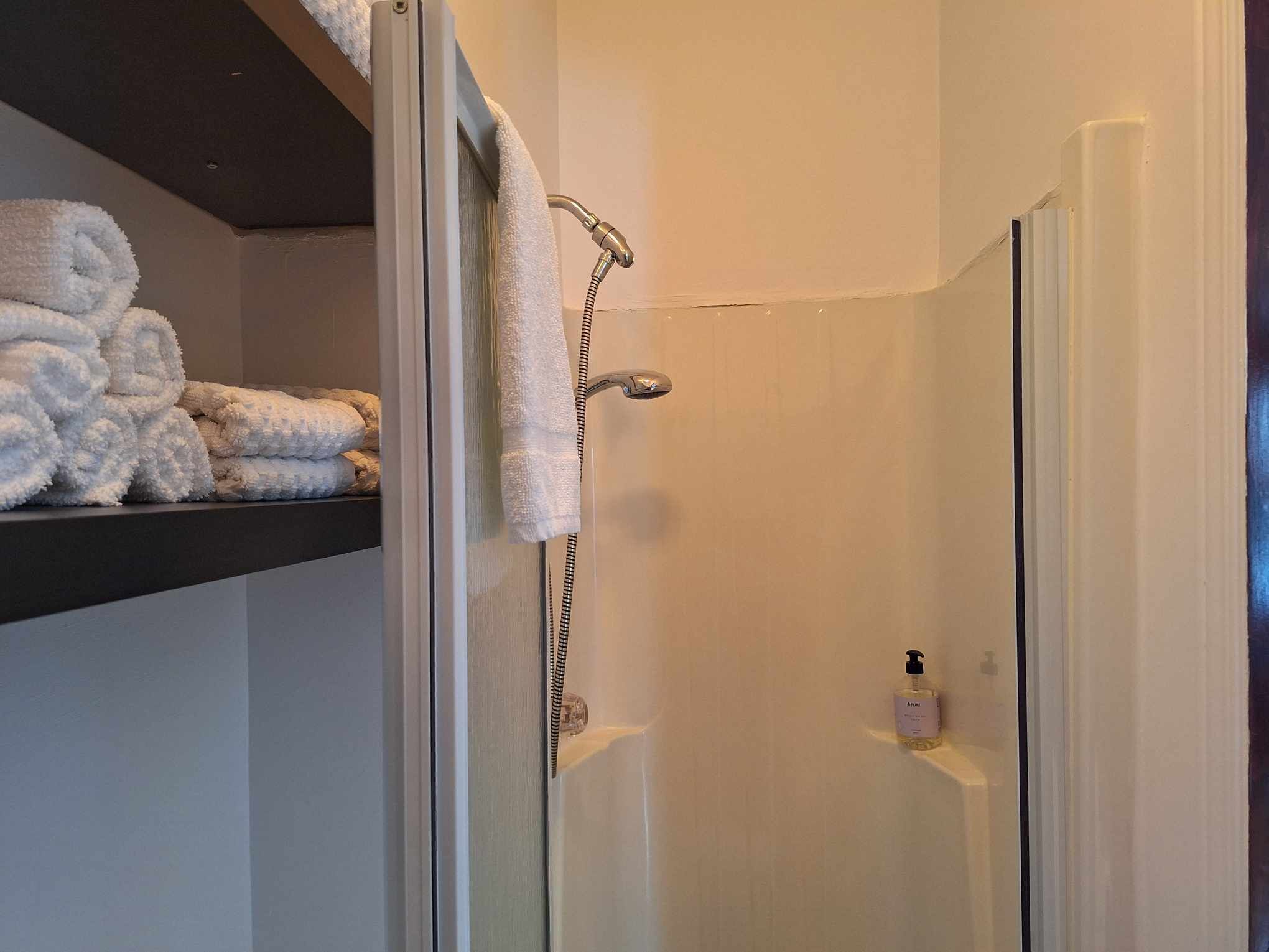 A shower with a glass door, a towel rack with folded towels, and a bottle of shampoo on the wall.