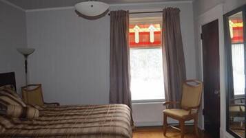 Ein Schlafzimmer mit Bett, Sessel, einem Fenster mit Vorhängen und einer Deckenleuchte.