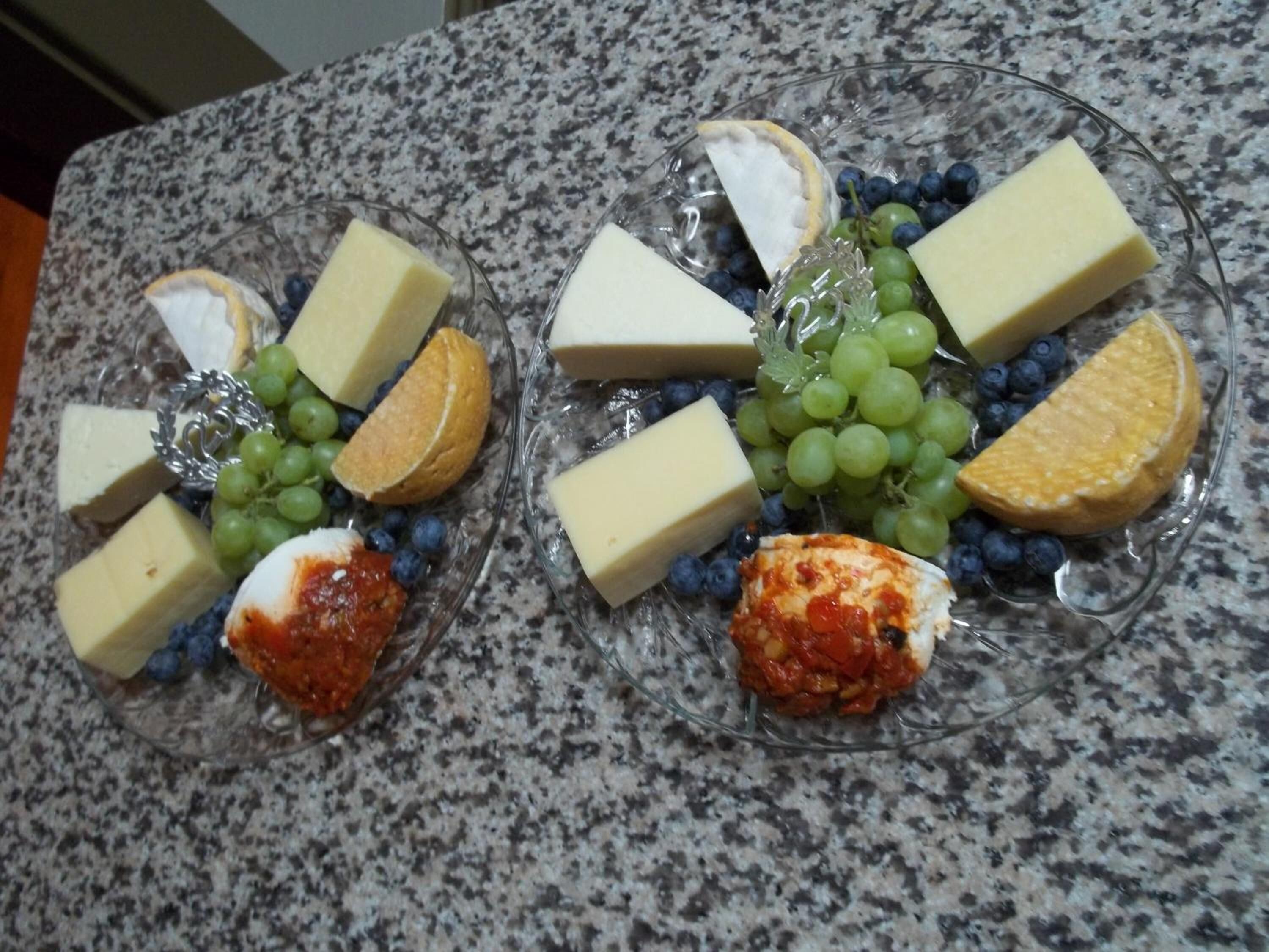 Two glass plates with cheese, grapes, and orange slices on a granite countertop.