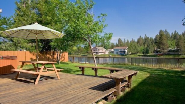A wooden deck with picnic tables and an umbrella, surrounded by a grassy yard and a lake view.
