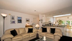 A living room with two beige sofas, a glass coffee table, and a kitchen area in the background.