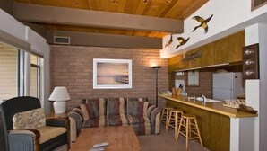 A cozy living area with a sofa, a wooden coffee table, a lamp, and a kitchen counter with stools.