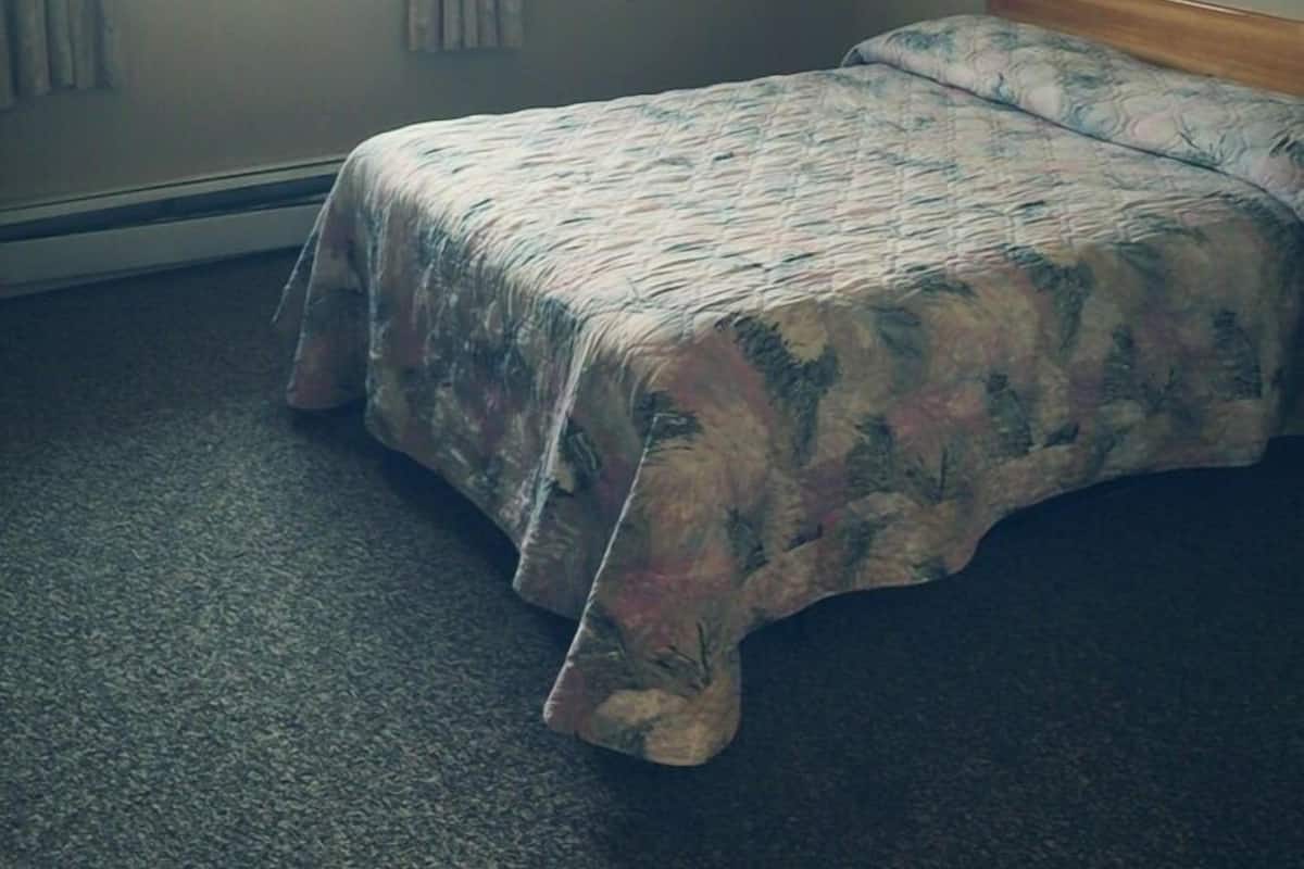 Une petite chambre avec un lit simple, une fenêtre, un tableau encadré au mur et un sol recouvert de moquette.