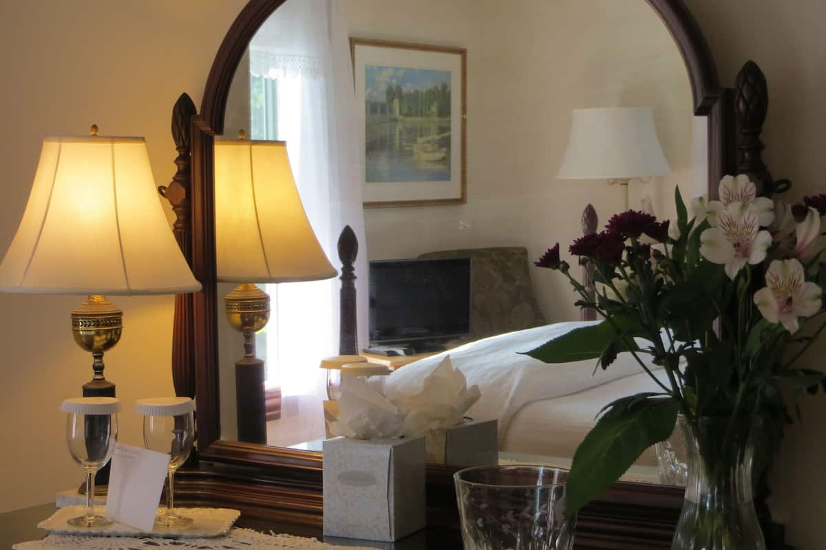 A hotel room with a bed, two lamps, a vase with flowers, a bedside table with a tissue box, and a mirror reflecting a painting.