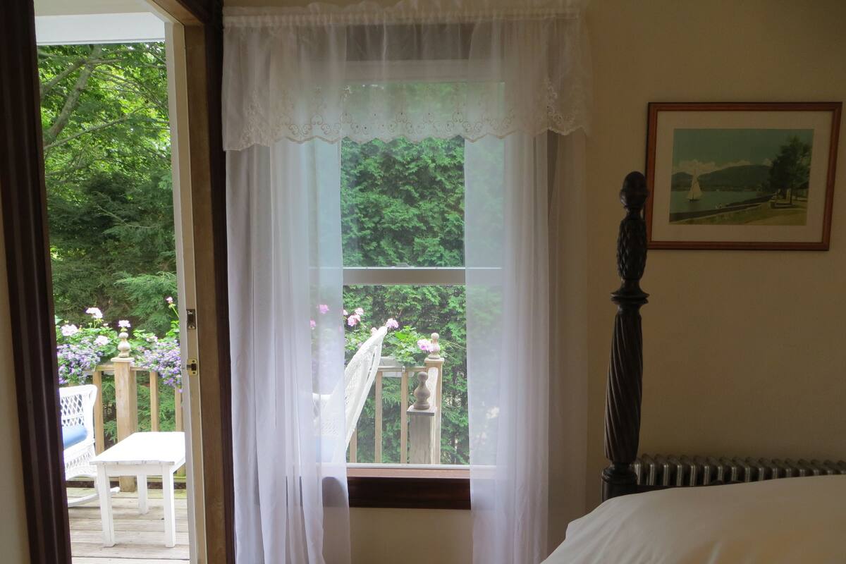 A bedroom with a large window, a bed, a framed picture on the wall, and a view of a garden outside.