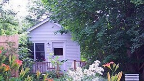 A small white house with a porch, surrounded by lush greenery and flowering plants. - Atlantic Ark Inn (Boothbay Harbor)