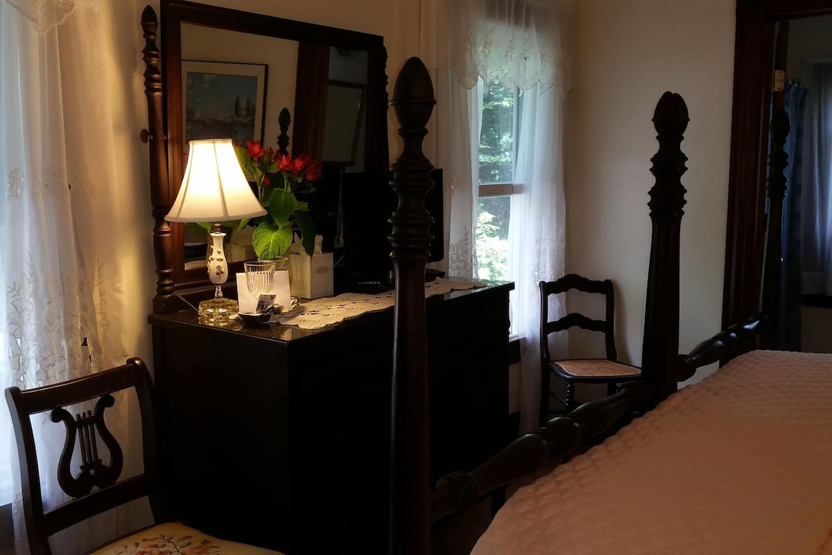 A bedroom with a bed, a wooden chair, a dresser with a mirror, a lamp, and a vase with flowers.
