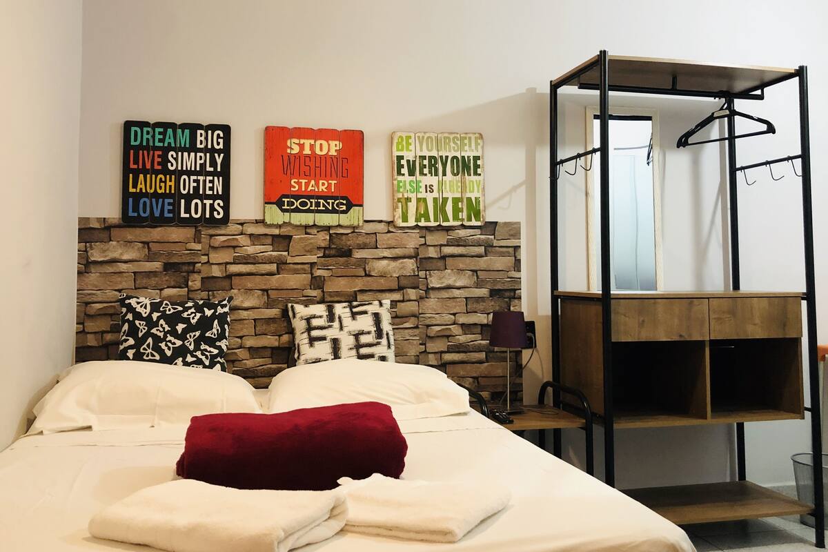 Ein ordentlich gemachtes Bett mit einem roten Kopfkissen, eine Steinwand dahinter und motivierende Schilder an der Wand.