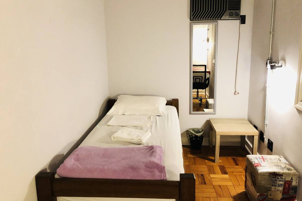 Ein kleines Zimmer mit einem Einzelbett, einem Holzrahmen, weißer Bettwäsche, einem kleinen Tisch, einem Spiegel und einem Heizkörper.