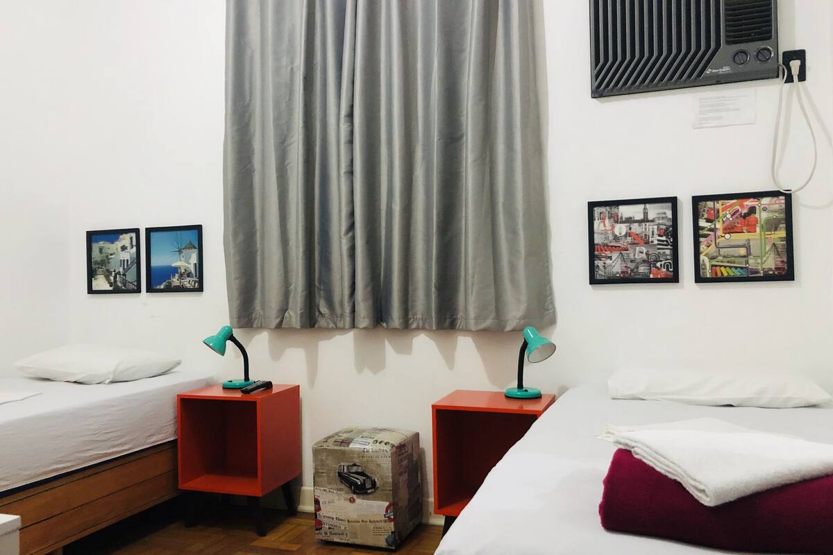 Ein Zimmer mit zwei Einzelbetten, einem grauen Vorhang, zwei an der Wand befestigten Bilderrahmen, einem kleinen Heizgerät und einer Kiste auf dem Boden.
