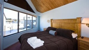 A bedroom with a large bed, wooden headboard, a nightstand, a lamp, and a window with a view of a snowy landscape.