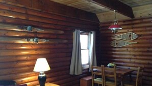 A cozy cabin interior with wooden walls, a dining table with chairs, a lamp, and a window with curtains.