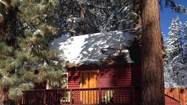 A cabin with a wooden deck and a snow-covered roof, surrounded by trees.