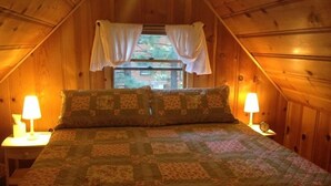 A cozy, wooden-paneled room with a bed, two bedside lamps, and a window with curtains.