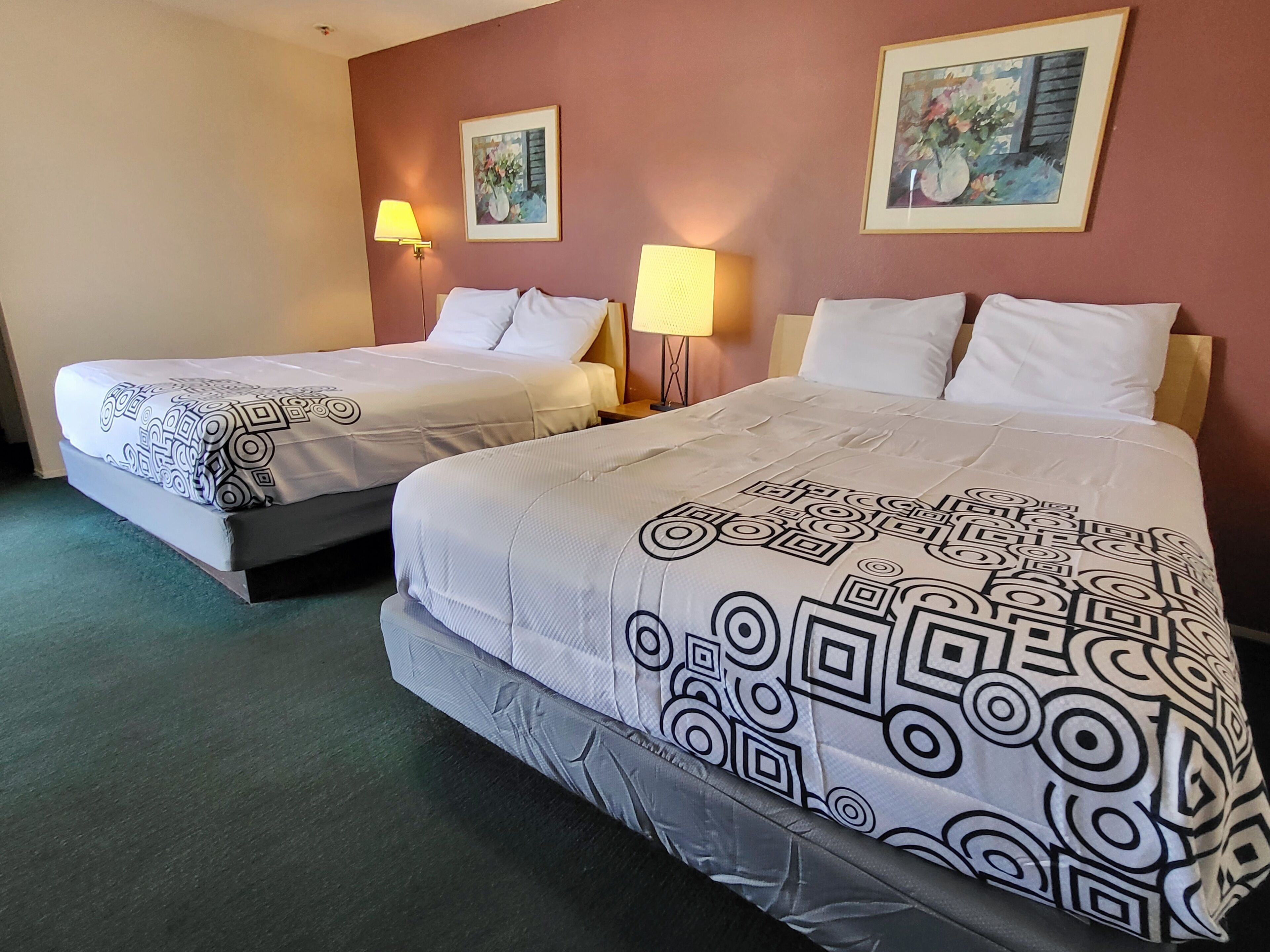 A hotel room with two beds, each with a patterned bedspread and white pillows. There are two wall-mounted lamps and two framed artworks on the walls.