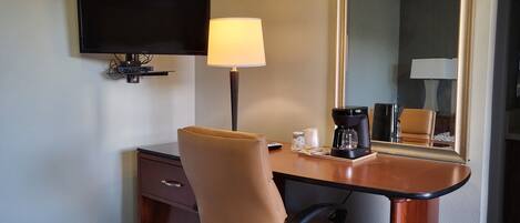 A hotel room with a desk, chair, television, lamp, coffee maker, and a large mirror.
