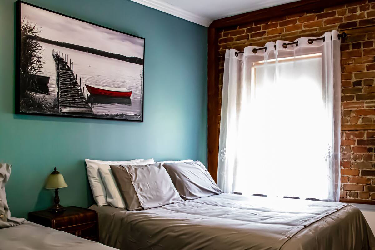 Un dormitorio con cama, mesita de noche, lámpara, un cuadro y una pared de ladrillo.