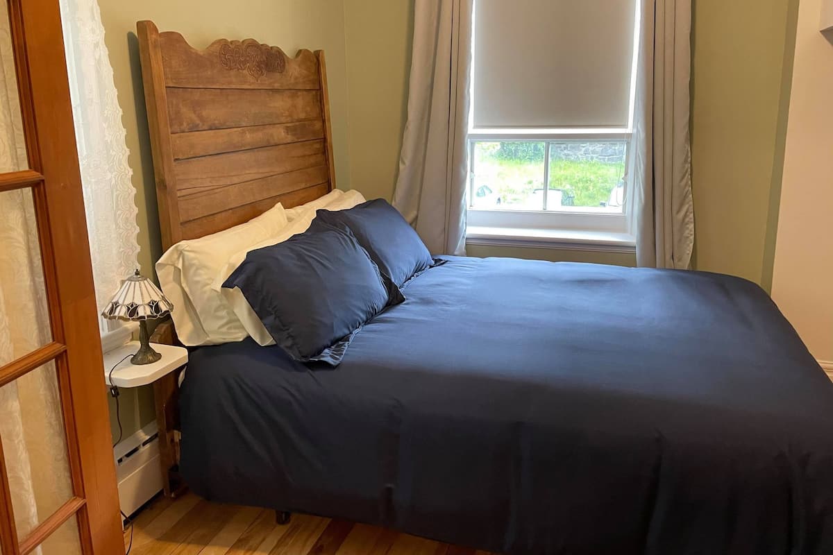 Un dormitorio con cabecera de madera, una mesita de noche blanca con una lámpara y una ventana con cortinas.