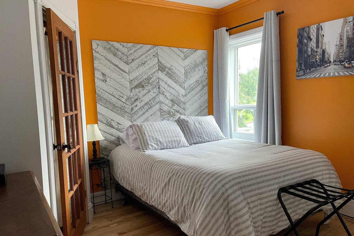 Un dormitorio con una cama, una puerta de madera, un ventanal con cortinas y un cuadro de un paisaje urbano en la pared.