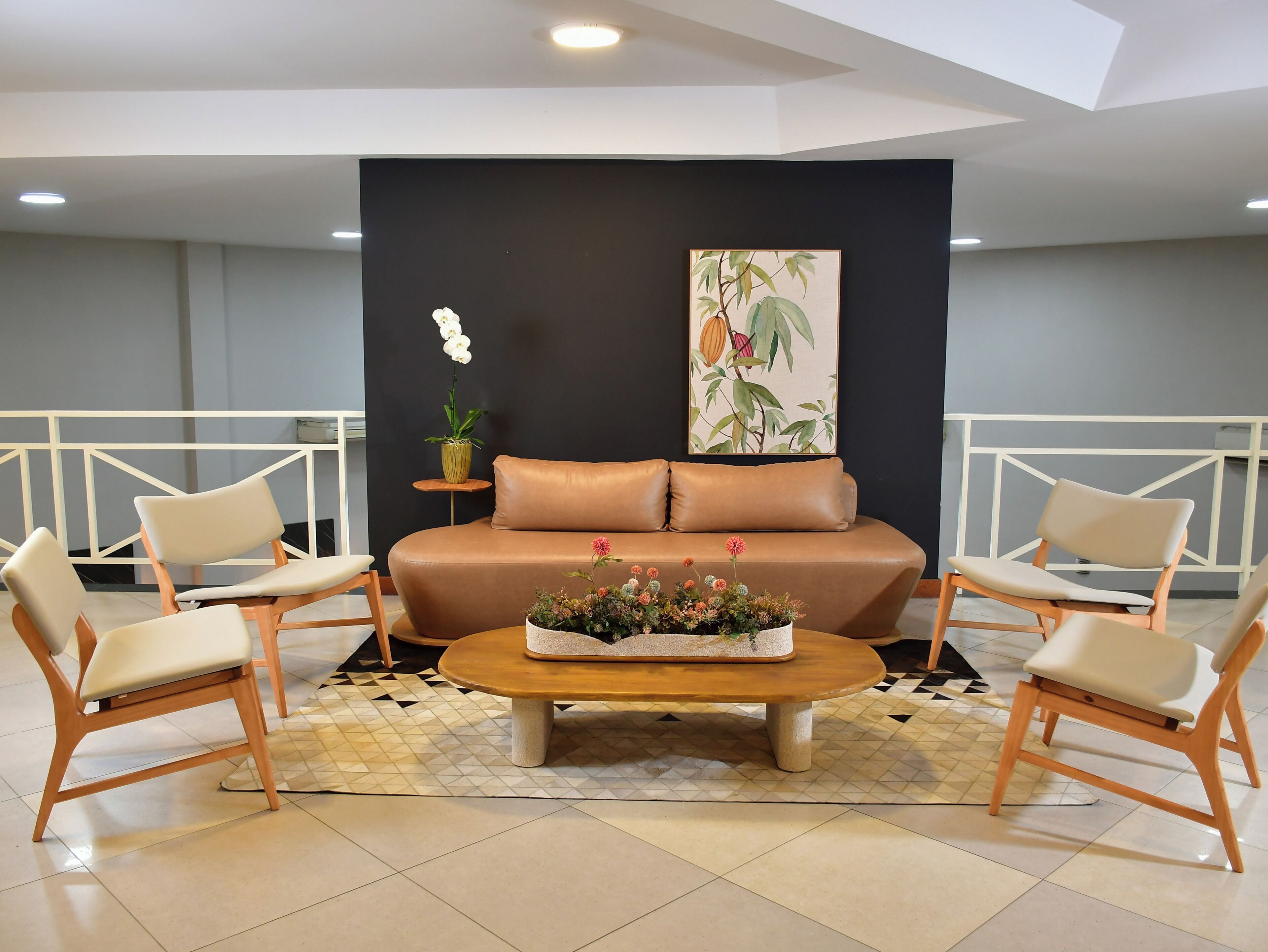 Uma sala de estar moderna com um sofá, duas poltronas, uma mesa de centro com flores, uma planta em vaso e uma parede com uma impressão botânica.