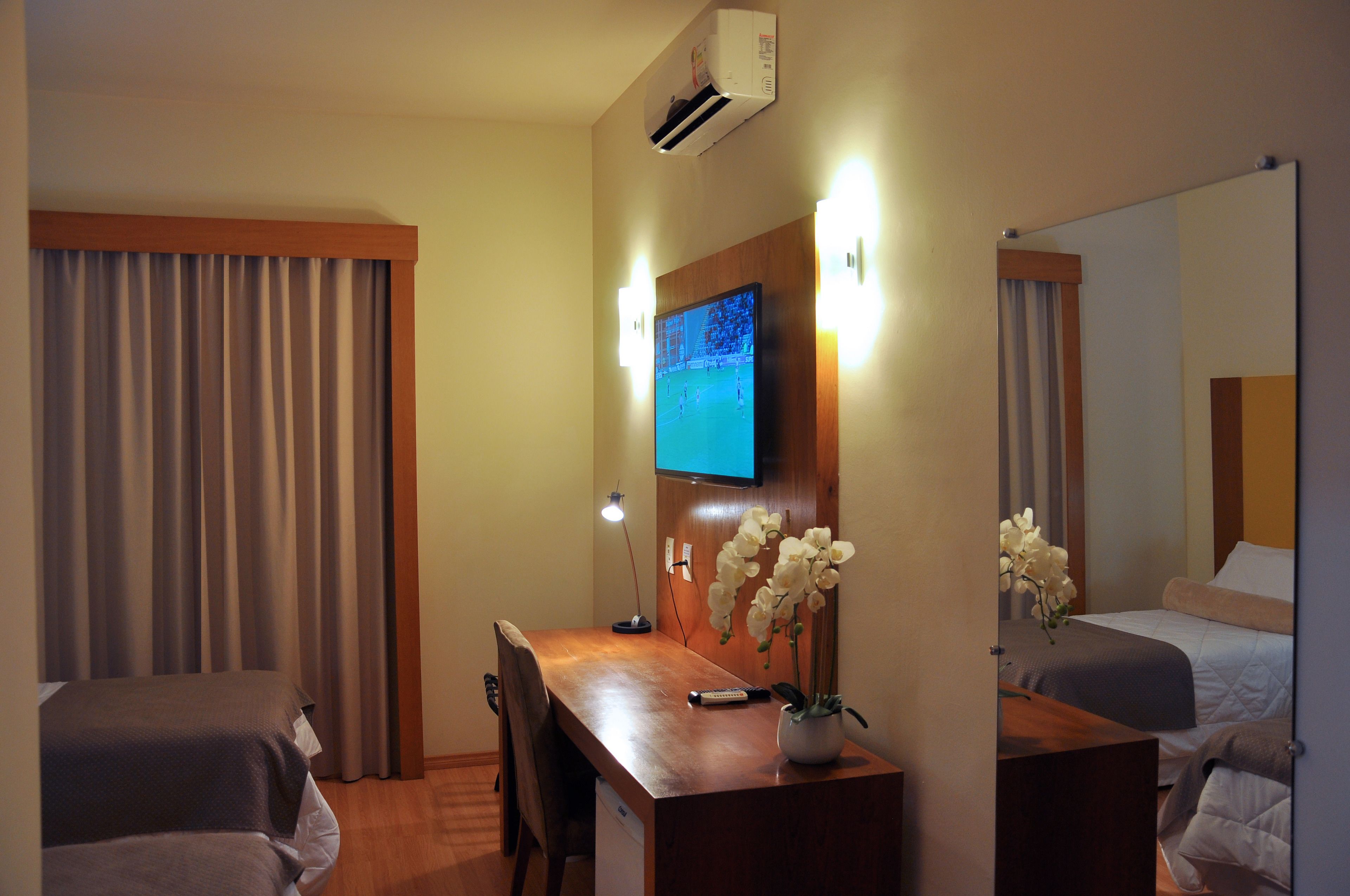 Quarto de hotel com uma escrivaninha de madeira, televisão fixada na parede, cama com cabeceira e uma orquídea em vaso.