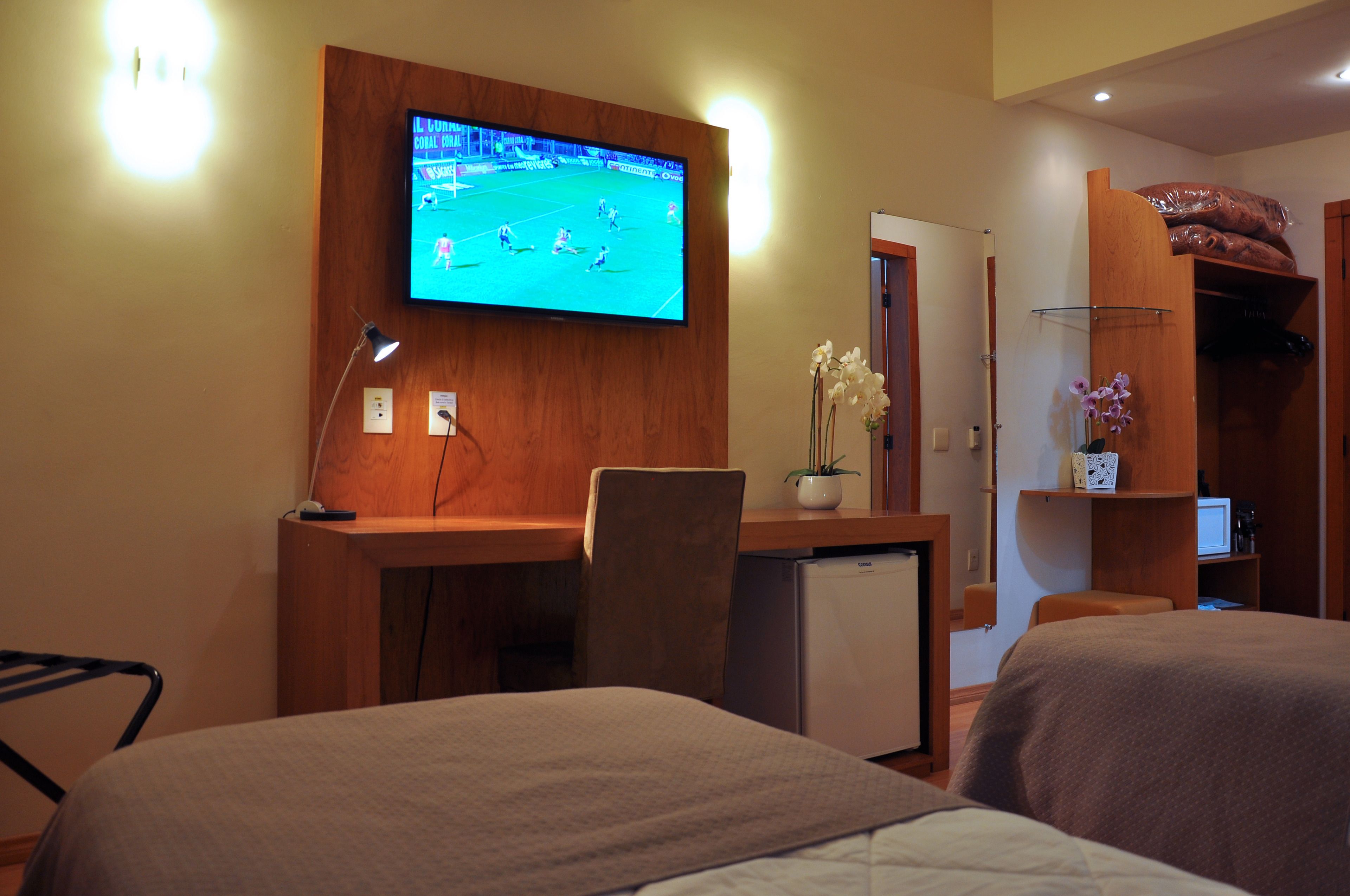 Habitación de hotel con televisión montada en la pared, escritorio con silla, refrigerador pequeño y cama con cabecera.