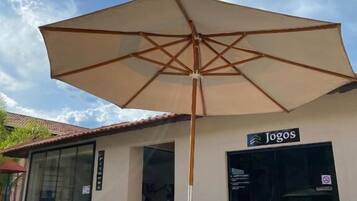 Un área de estar al aire libre con una mesa, sillas y un paraguas grande, junto a un edificio con un letrero que dice "Jogos".