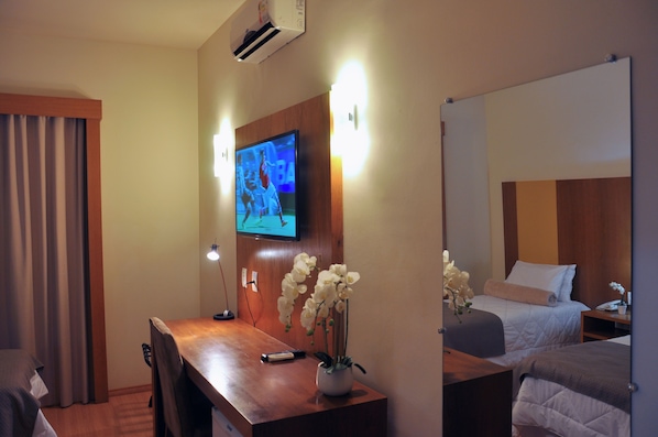 A hotel room with a wooden desk, a television mounted on the wall, a bed with a white comforter, and a vase of white flowers. - Giorni Hotel Boulevard (Resende)