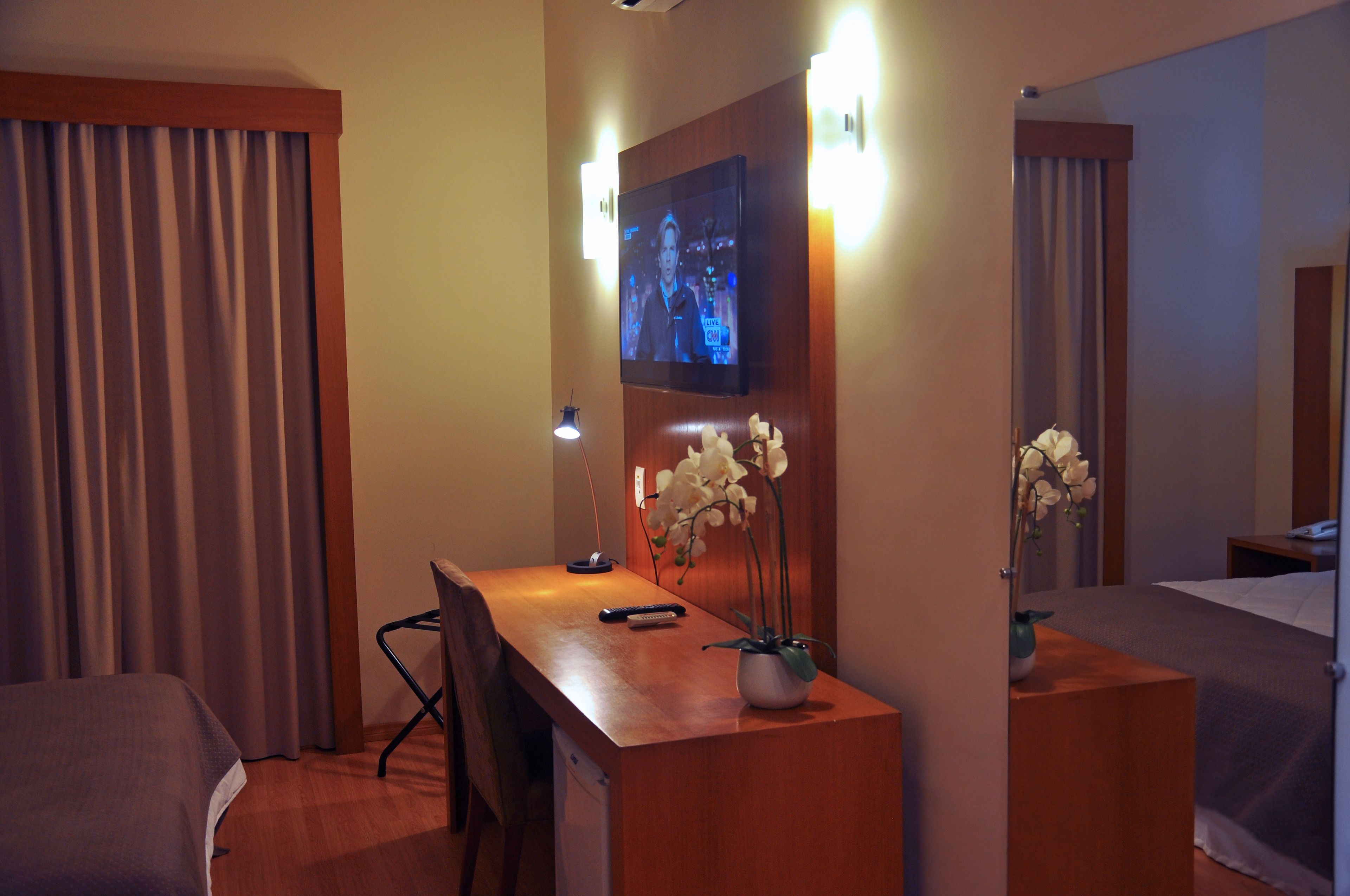 Een hotelkamer met een houten bureau, een aan de muur bevestigde televisie, een bed en een spiegel.