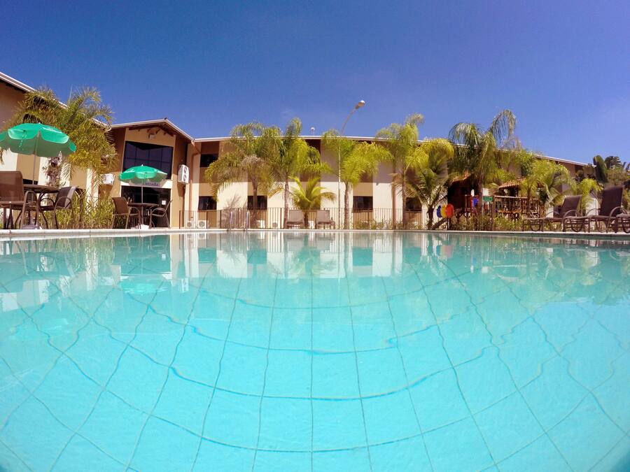 Uma piscina azul límpida com bordas azulejadas, rodeada por espreguiçadeiras e guarda-sóis, com um edifício com várias varandas e palmeiras ao fundo.