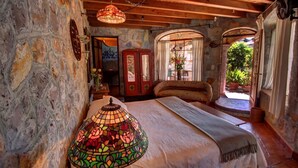 A rustic bedroom with a stone wall, a bed with a white bedspread, a stained glass lamp, and a red cabinet. - Hotel Casa Angelitos (San Miguel de Allende)