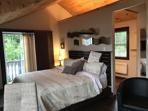 Ein Schlafzimmer mit Bett, Nachttisch, Lampe, Fenster und Blick auf Grünlandschaft.