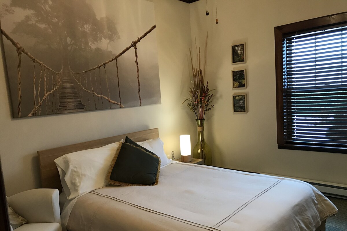 Ein ordentlich bezogenes Bett mit Kopfteil, ein Nachttisch mit Lampe, eine Vase mit Trockenblumen und ein Fenster mit Jalousien.
