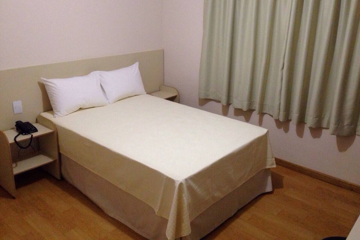 Una habitación de hotel sencilla con una cama, una mesita de noche con un teléfono y una ventana con cortinas.