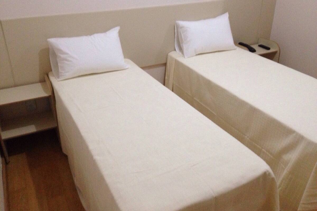 Habitación de hotel con dos camas individuales, cada una con una almohada blanca y una colcha blanca. Hay una mesita de noche de madera con un teléfono encima.