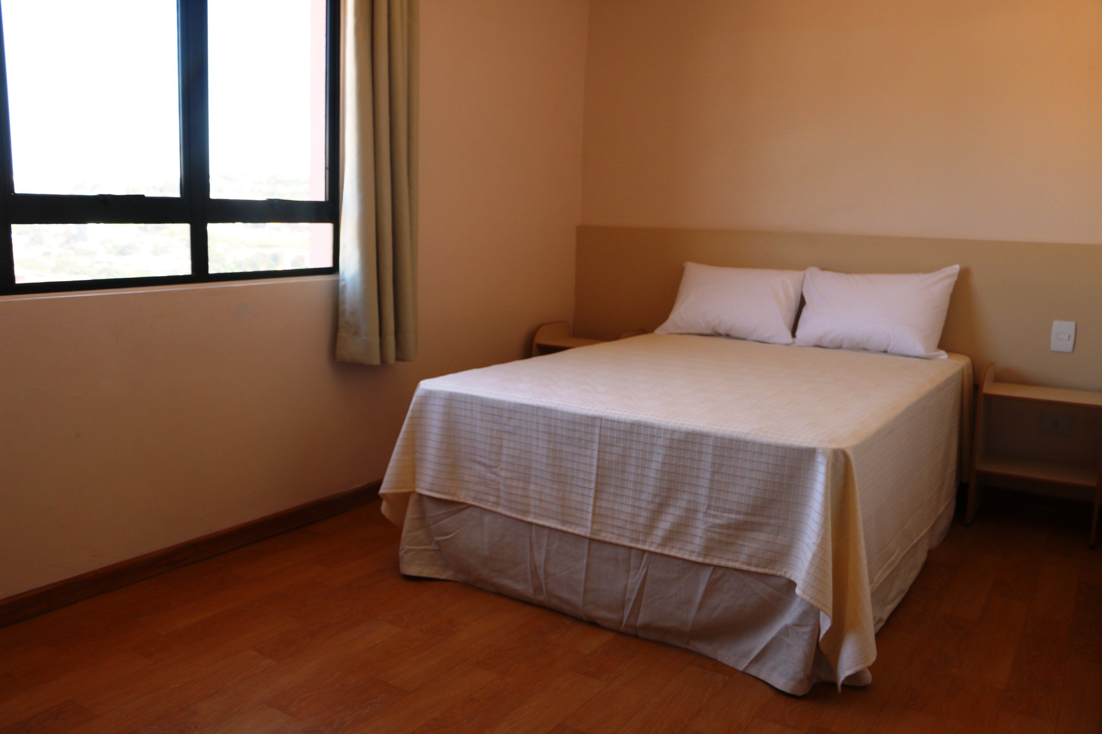 Una cama individual con una colcha blanca en una habitación con suelo de madera y una ventana con cortinas.