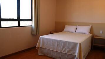 Una cama individual con una colcha blanca en una habitación con suelo de madera y una ventana con cortinas.