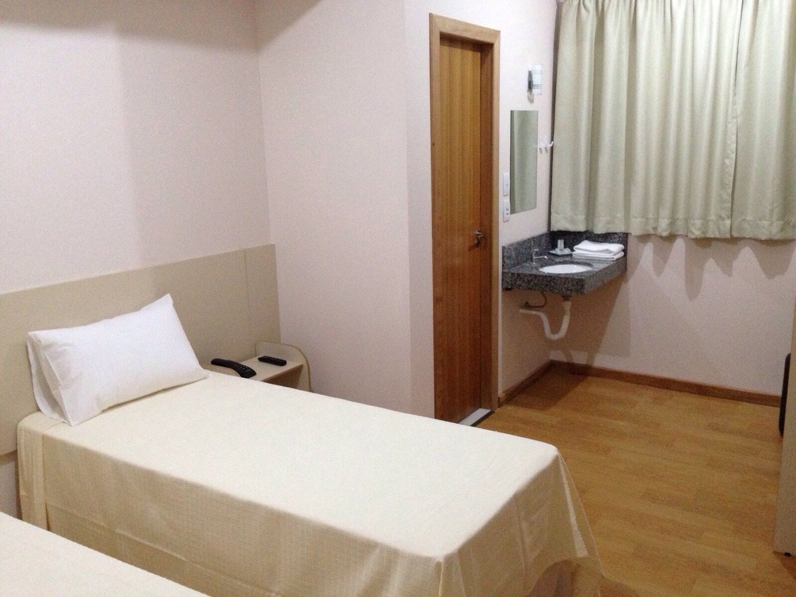 A small hotel room with a single bed, a bedside table, a small desk with a chair, a bathroom with a sink and mirror, and a wooden floor.
