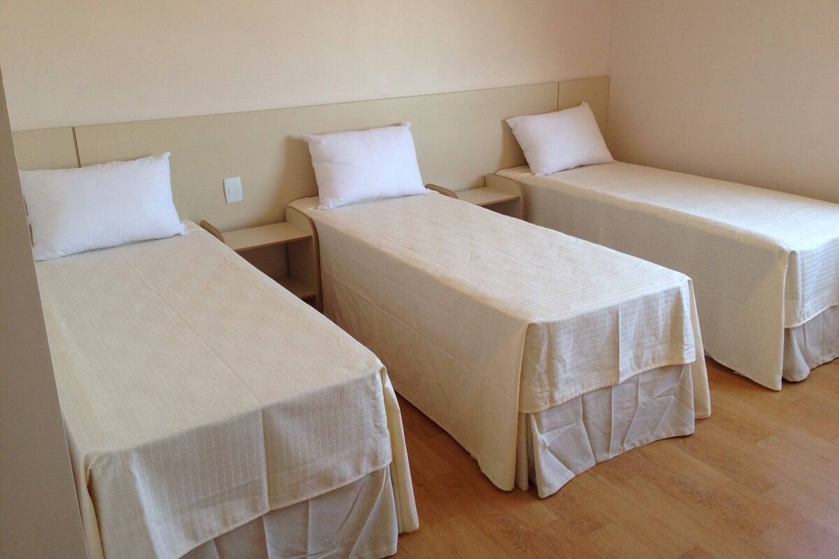 Tres camas individuales con sábanas blancas en una habitación con suelo de madera y paredes color beige.