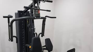 Un gimnasio pequeño y funcional con una estación de entrenamiento multifuncional, que incluye un rack para sentadillas, prensa de banca y varios discos de pesas.