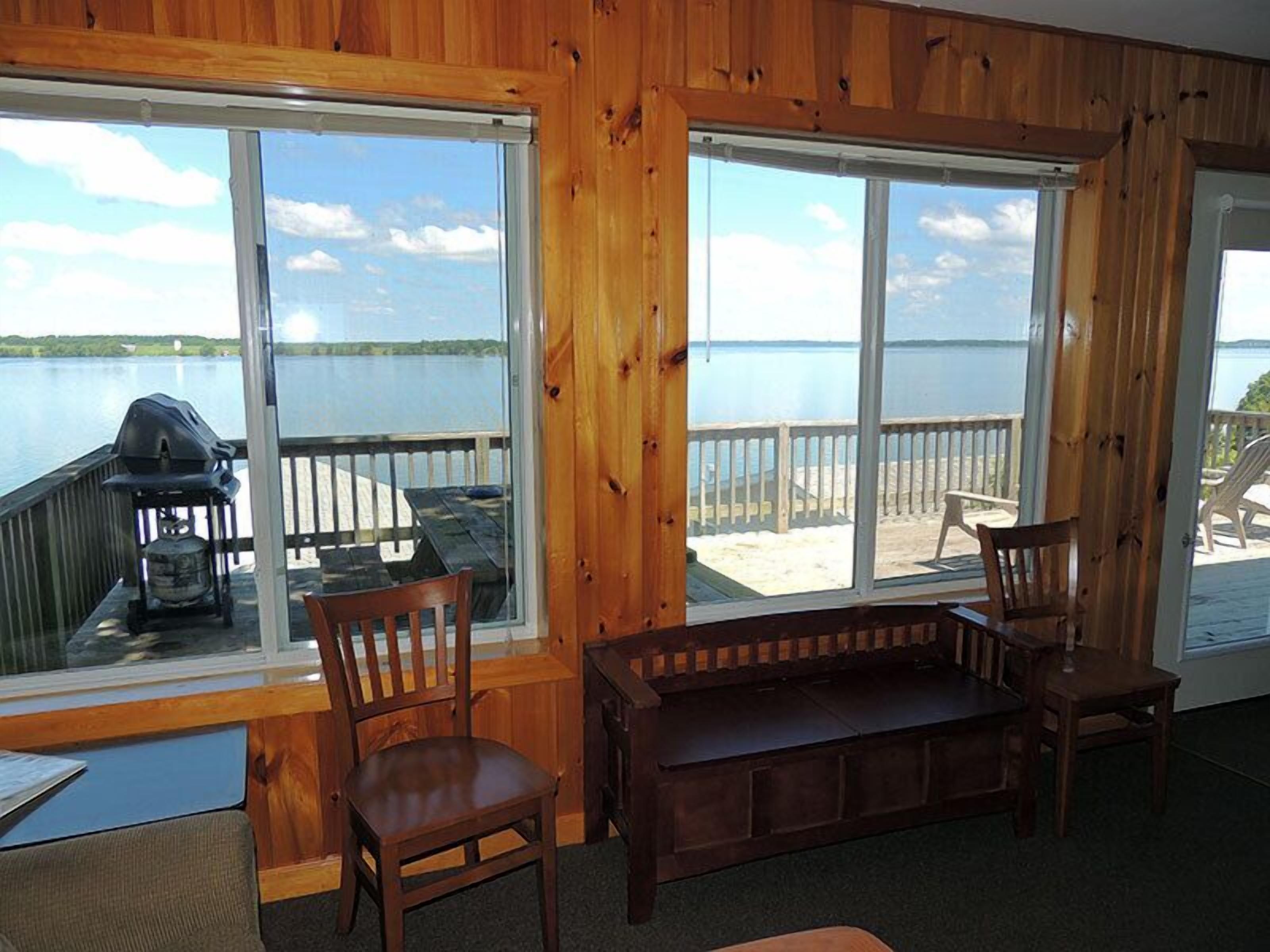 A wooden cabin with large windows overlooking a lake, a barbecue grill, and a wooden bench.