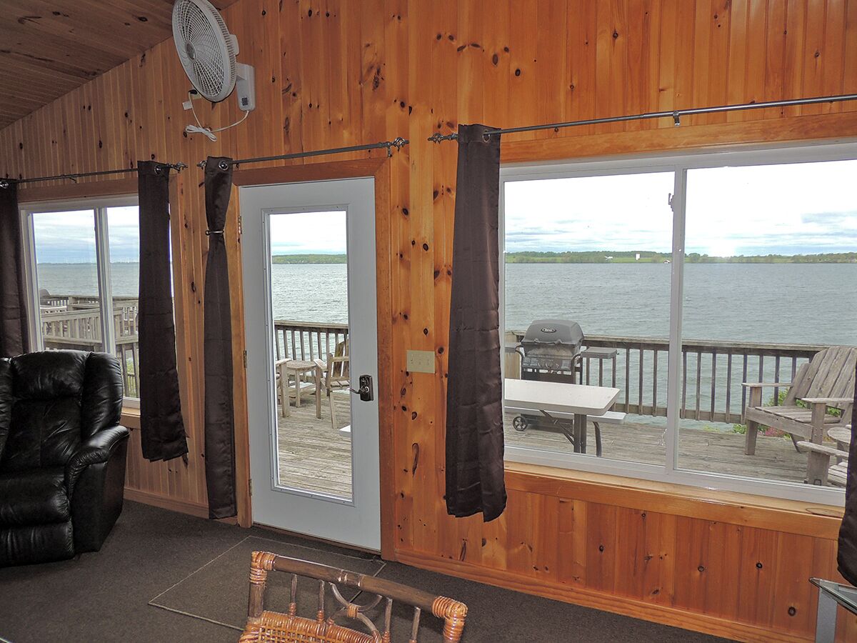 Une cabine avec un intérieur en bois, une porte donnant sur une terrasse avec vue sur l’eau, un ventilateur et une chaise.