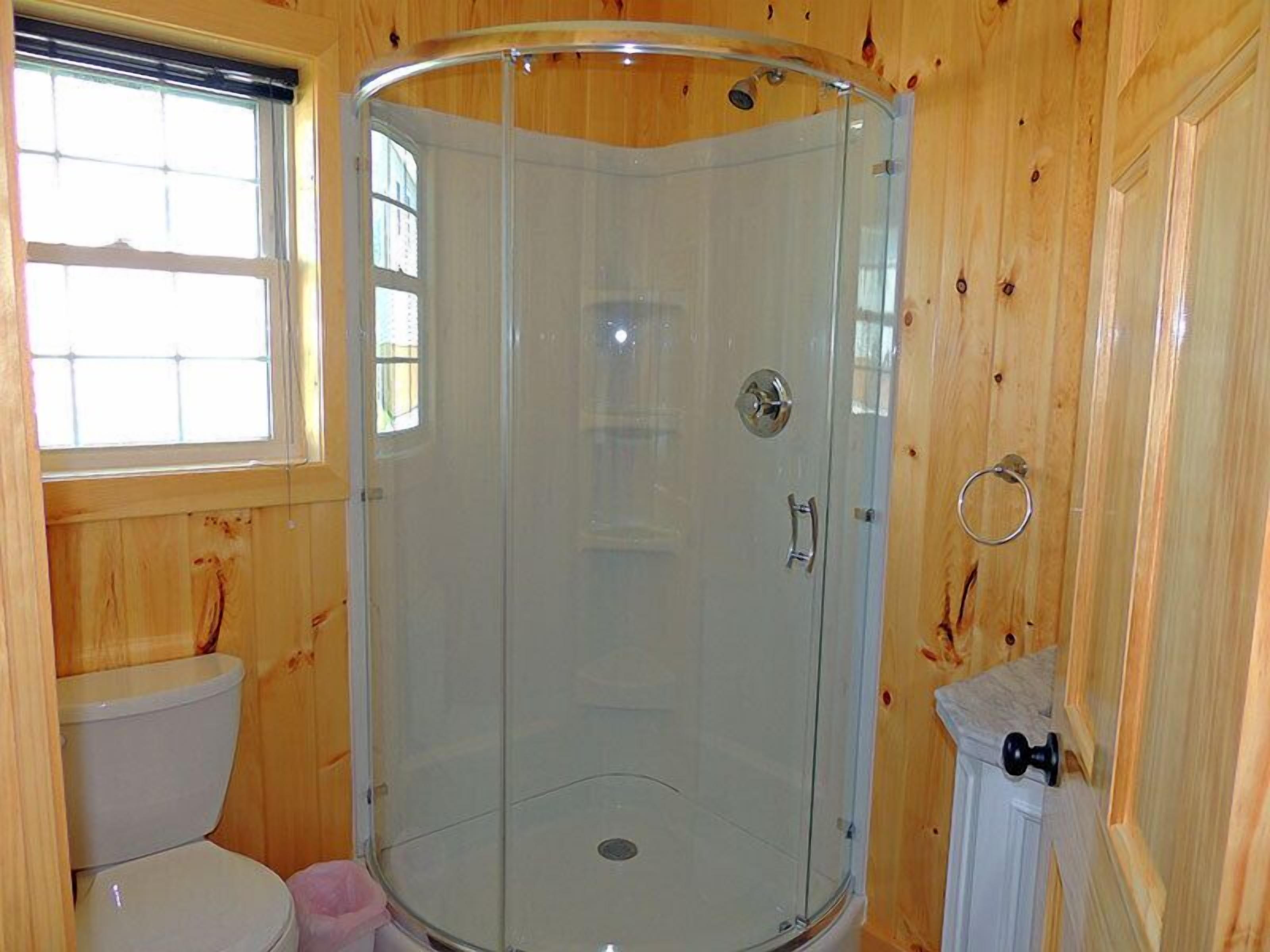 A wooden cabin bathroom with a glass shower enclosure, a toilet, and a window.