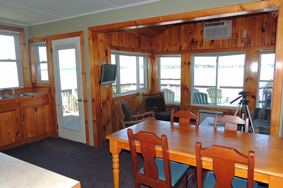 Interior de una cabaña de madera con una mesa de comedor, sillas, un televisor y un balcón con sillones.