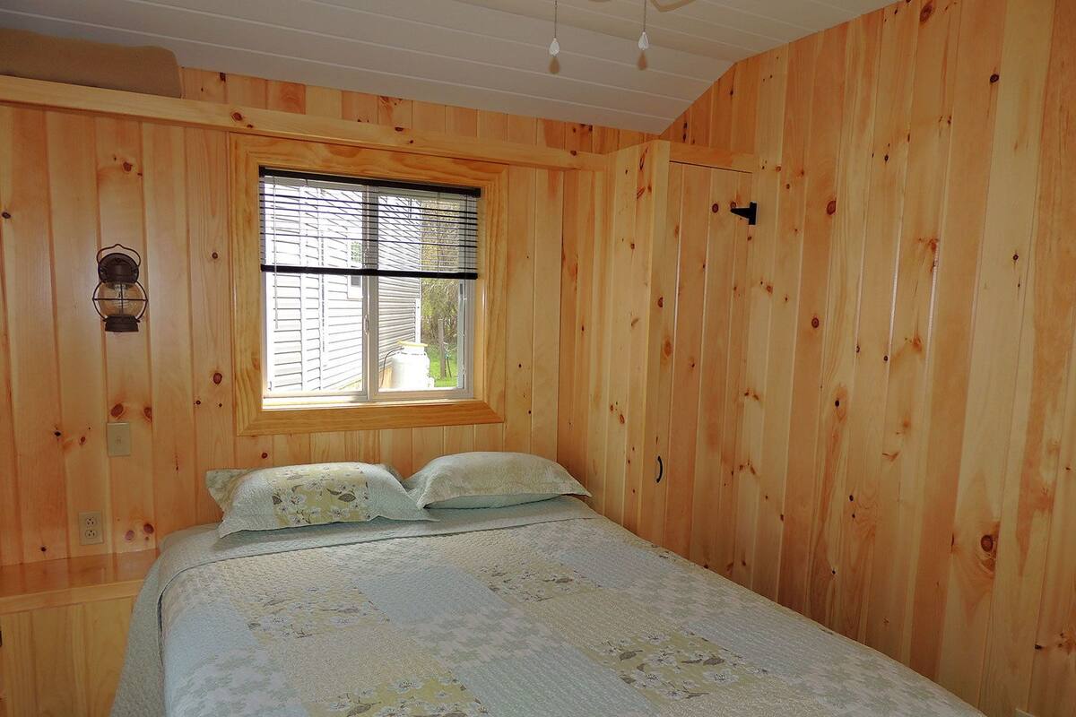 Habitación con paneles de madera, una cama, una ventana y una lámpara fijada a la pared.
