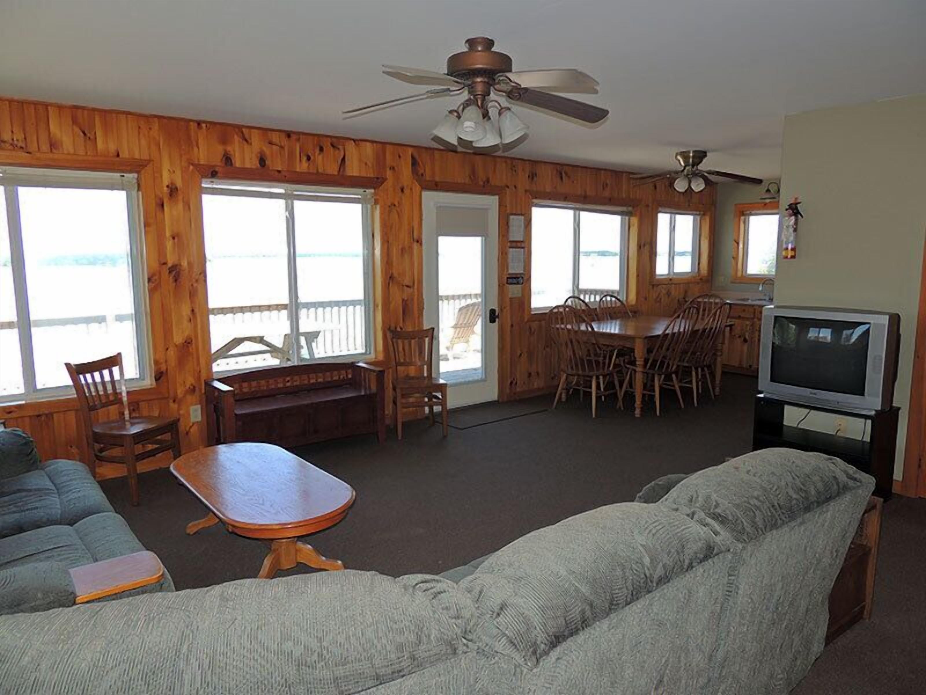 A cozy cabin interior with a wooden interior, a dining table with chairs, a sofa, a television, and large windows offering a view.
