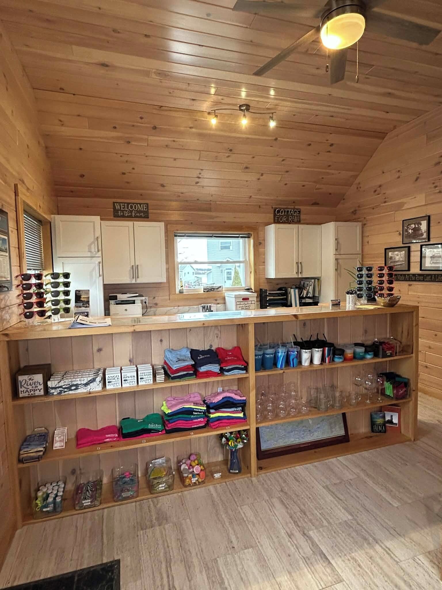Une chambre confortable, aménagée dans le style d’un chalet, avec des murs et un plafond en bois. Elle comprend un espace d’accueil avec une pancarte « Bienvenue », une pancarte « Chalet à louer » et des étagères remplies de divers articles, notamment des vêtements pliés, des collations et des verres.
