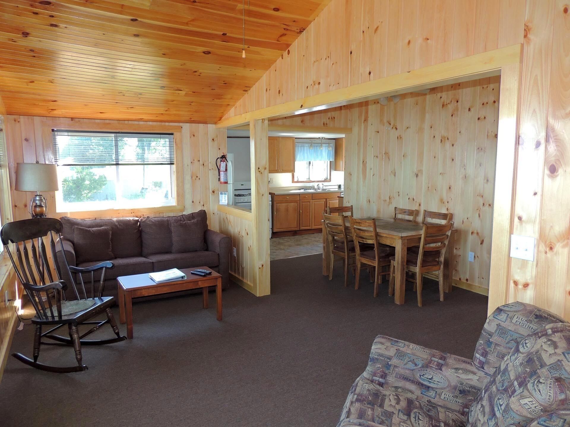 A wooden interior with a sofa, a rocking chair, a dining table with chairs, and a kitchen area.
