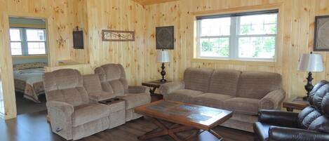A cozy cabin interior with wooden walls, a large window, a sofa, a coffee table, and a leather chair.